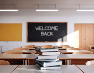 A classroom, with welcome back on the board and a stack of books on the desk