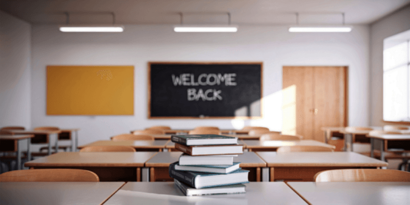 A classroom, with welcome back on the board and a stack of books on the desk