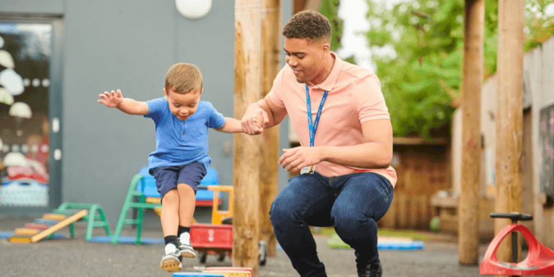 Teaching assistant helping a child climb on outdoor playground equipment